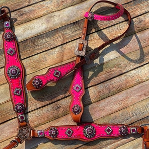 May include: A vibrant pink and brown horse tack set. The set includes a headstall, breast collar, and reins, all adorned with silver-toned studs and sparkling pink gemstones. The leather is brown, and the pink material has a textured pattern.