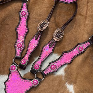 Könnte beinhalten: Ein Pferdegeschirr-Set aus dunkelbraunem Leder mit leuchtend pinkfarbenen Leopardenmuster-Applikationen. Das Set besteht aus einem Kopfstück und einem Vorderzeug, verziert mit silbernen Nieten, runden Blumen-Conchos und kupferfarbenen Schnallen.
