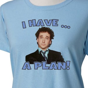 Pode incluir: Uma camiseta azul claro com uma imagem de um homem de terno e gravata. O texto "I HAVE... A PLAN!" está impresso na camiseta.