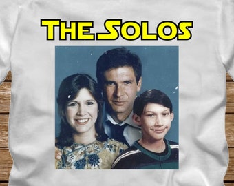May include: White t-shirt with a photo of Han Solo, Princess Leia, and a young boy. The text "THE SOLOS" is above the photo and "WHITE" is below the photo.