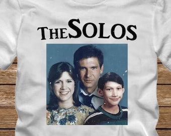 May include: White t-shirt with a black graphic of a family photo. The text "THE SOLOS" is above the photo. The word "WHITE" is printed in large white letters at the bottom of the shirt.