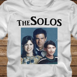 May include: White t-shirt with a black graphic of a family photo. The text "THE SOLOS" is above the photo. The word "WHITE" is printed in large white letters at the bottom of the shirt.