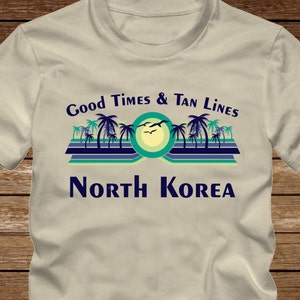 May include: A beige t-shirt with a graphic design featuring palm trees, a sun, and the text "Good Times & Tan Lines North Korea".