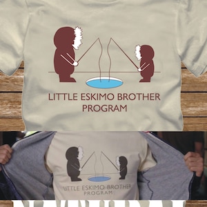 Può includere: Una maglietta beige con una grafica di due figure stilizzate che pescano sul ghiaccio. Il testo "Little Eskimo Brother Program" è stampato sotto la grafica.