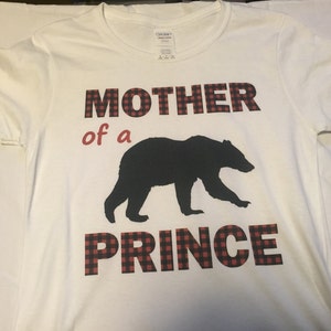 Op de afbeelding: Wit T-shirt met rood-zwarte geruite tekst die "Moeder van een Prins" luidt. Een zwart silhouet van een beer bevindt zich tussen de woorden "van een" en "Prins".