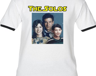 May include: A white t-shirt with a black ringer, featuring a photo of Han Solo, Princess Leia, and a young boy with the text "THE SOLOS" in yellow above the photo.