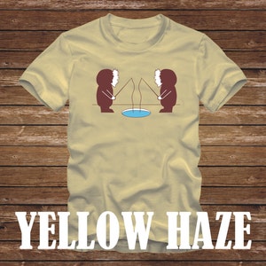 Può includere: Una maglietta beige con una grafica di due personaggi dei cartoni animati che indossano cappelli invernali e tengono canne da pesca. I personaggi si fronteggiano e pescano in un piccolo lago blu. Il testo "YELLOW HAZE" è stampato in bianco nella parte inferiore della maglietta.