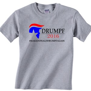 Op de afbeelding: Grijs T-shirt met een blauwe en rode afbeelding van een gezicht met een rood en blauw kapsel. De tekst "DRUMPF 2016 #MAKEDONALDDRUMPFAGAIN" staat op het T-shirt gedrukt.