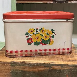 May include: A rectangular metal storage tin with a bright orange lid and a cream-colored body. The front features a central floral illustration of yellow and red flowers with green leaves, accented by a red and white checkered border at the base.
