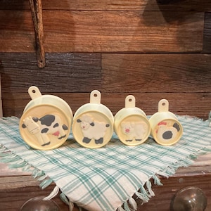 May include: A set of four off-white measuring cups with farm animal designs. Each cup features a different animal: a cow, a sheep, a pig, and a chicken. The cups are arranged on a green and white checkered cloth.