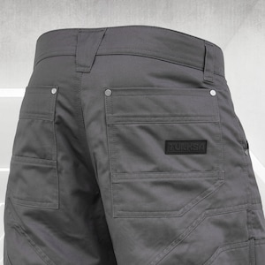 May include: Close-up of gray work pants with multiple pockets and reinforced stitching. The pants feature a black label with the text "TULKSA". The design includes decorative stitching and metal rivets.