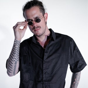 Puede incluir: Un hombre con una camisa negra de manga corta con botones y un tatuaje geométrico en el brazo. Lleva gafas de sol redondas con montura dorada y tiene barba.