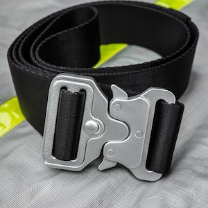 Könnte beinhalten: Ein schwarzer Nylon-Gürtel mit einer silbernen Metallschnalle. Die Schnalle hat ein einzigartiges Design mit einem kleinen Knopf an der Seite.