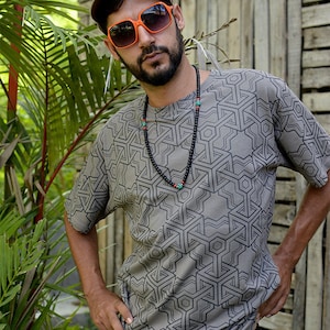 Peut inclure: Un homme portant un t-shirt gris à motif géométrique, une casquette marron et des lunettes de soleil orange. Il porte un collier de perles noires avec des accents turquoise.