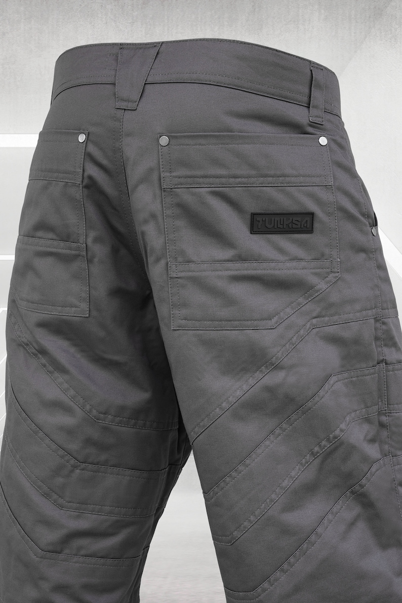 May include: Close-up of gray work pants with multiple pockets and reinforced stitching. The pants feature a black label with the text "TULKSA". The design includes decorative stitching and metal rivets.
