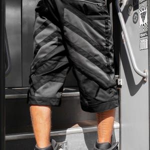 May include: Black cargo shorts featuring diagonal dark gray panels, a rear zippered pocket, and a small square patch on the waistband. The shorts have a structured design with reinforced stitching and a knee-length cut, providing a functional look.