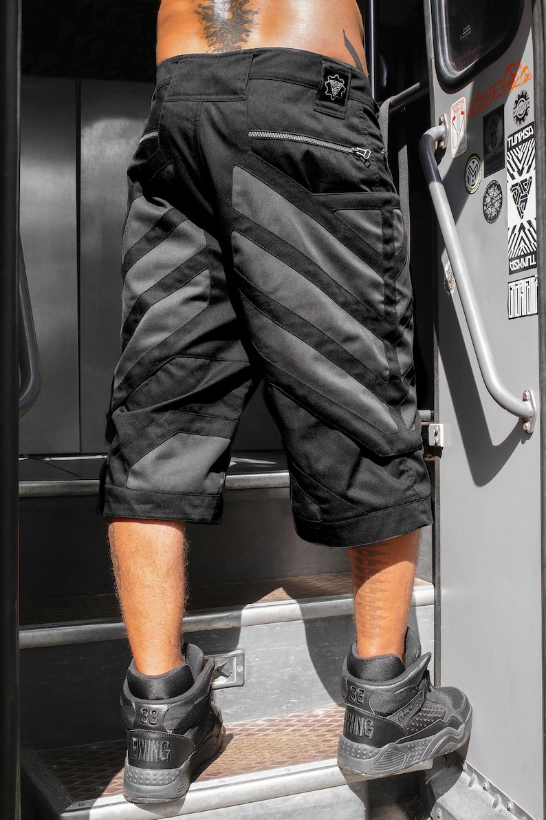 May include: Black cargo shorts featuring diagonal dark gray panels, a rear zippered pocket, and a small square patch on the waistband. The shorts have a structured design with reinforced stitching and a knee-length cut, providing a functional look.