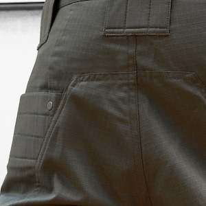 May include: Close-up of the back of a pair of dark green cargo pants with a large pocket on the left side and a smaller pocket on the right side. The pants have a belt loop and a zipper fly.
