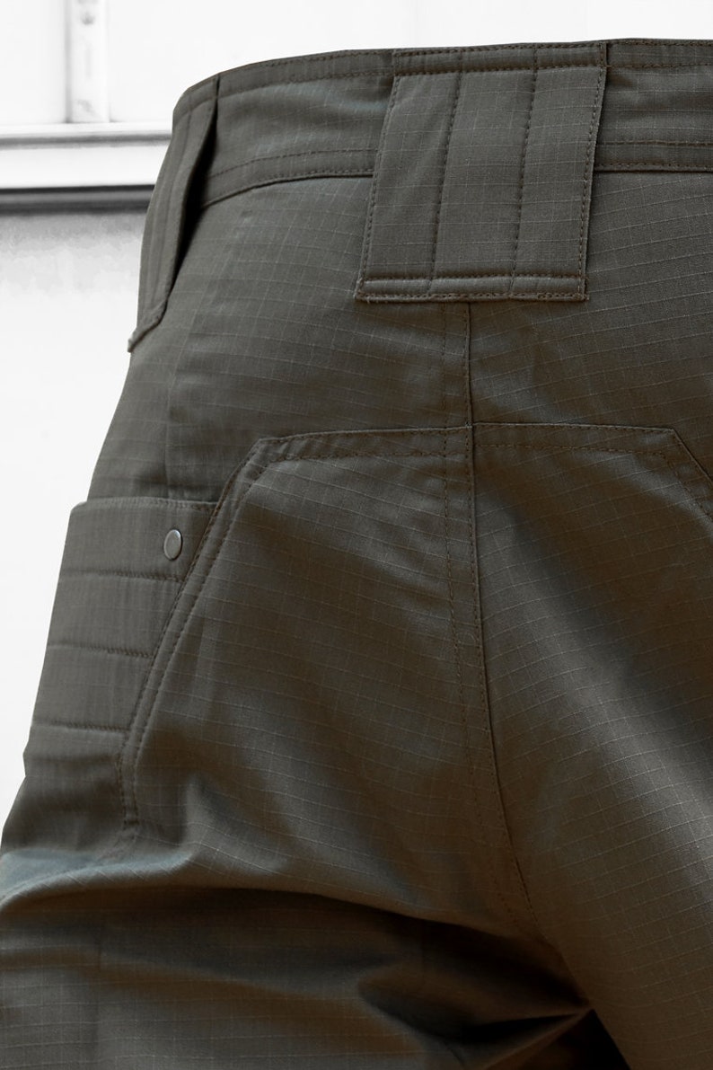 May include: Close-up of the back of a pair of dark green cargo pants with a large pocket on the left side and a smaller pocket on the right side. The pants have a belt loop and a zipper fly.