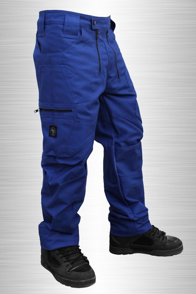 May include: Royal blue work trousers with multiple pockets and black zips. The trousers have a button and zip closure and are worn with black trainers. A patch is visible on the left leg.