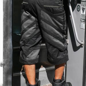 May include: Black cargo shorts featuring a diagonal panel design with alternating matte and dark grey fabric textures. The shorts include two horizontal zippered pockets on the back, a small rectangular patch on the waistband, and a loose knee-length fit.