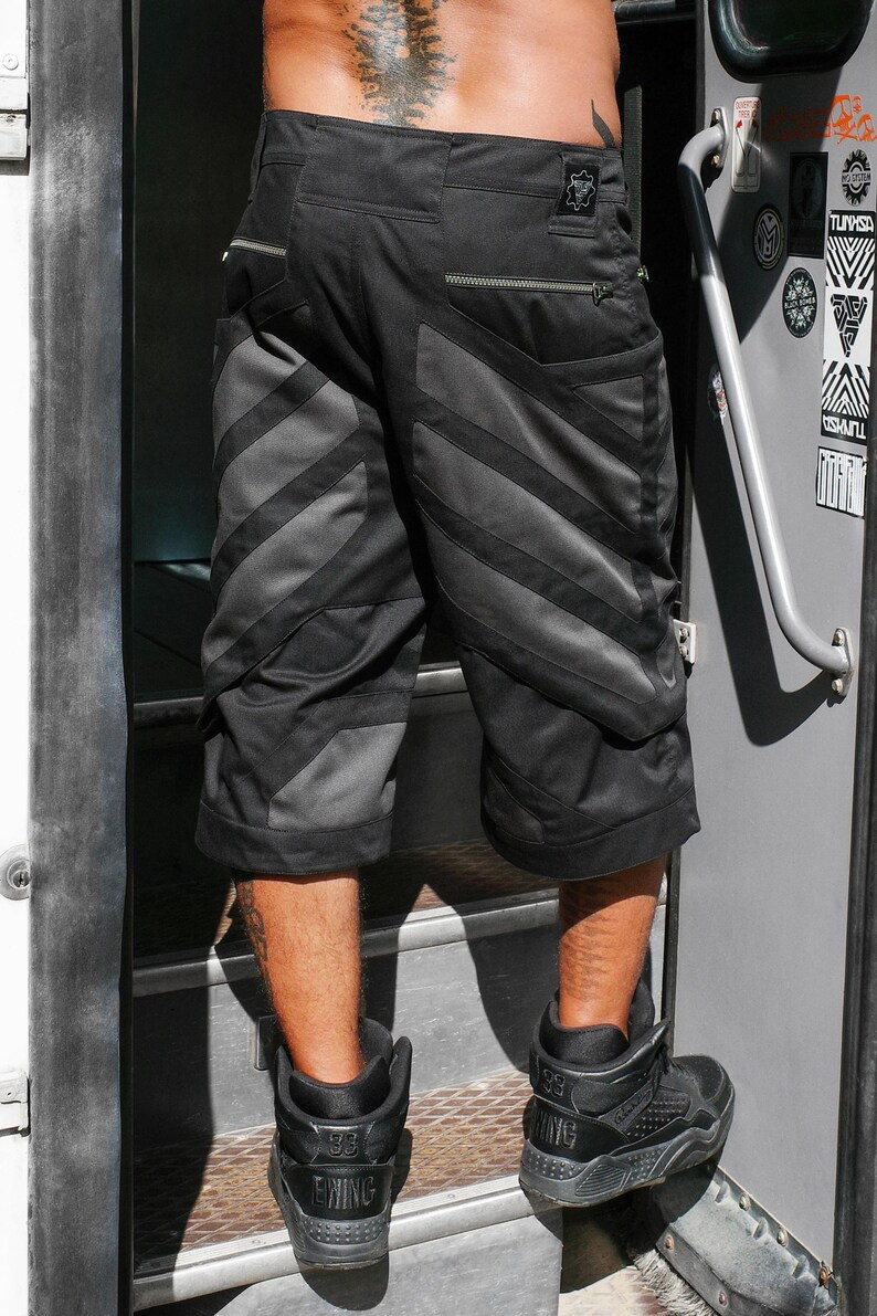 May include: Black cargo shorts featuring a diagonal panel design with alternating matte and dark grey fabric textures. The shorts include two horizontal zippered pockets on the back, a small rectangular patch on the waistband, and a loose knee-length fit.