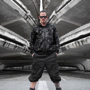 Könnte beinhalten: Ein Mann in einer schwarzen Kapuzenjacke mit geometrischem Muster, schwarzen Cargo-Shorts und schwarzen Stiefeln steht vor einer Betonstruktur.