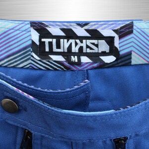 May include: Close-up of blue trousers with a decorative waistband featuring a geometric pattern in shades of purple, blue, and white. The label reads "TUNKS" in a bold, stylized font, with an "M" size indicator.