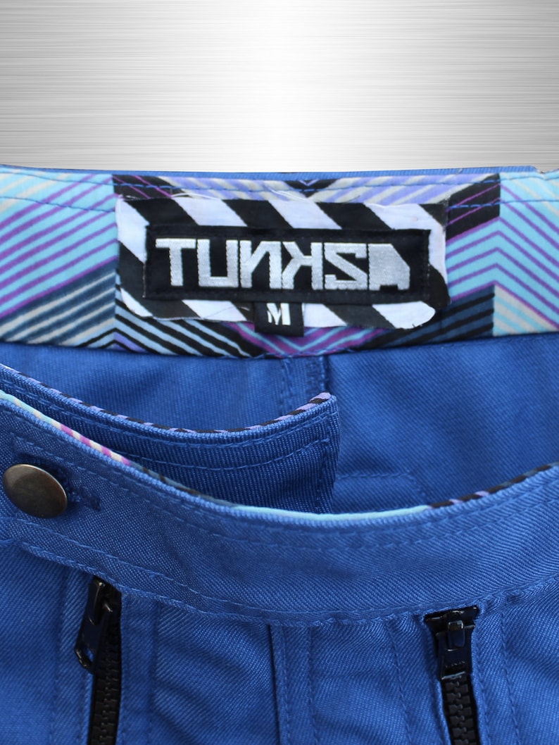 May include: Close-up of blue trousers with a decorative waistband featuring a geometric pattern in shades of purple, blue, and white. The label reads "TUNKS" in a bold, stylized font, with an "M" size indicator.
