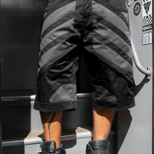 May include: Black shorts featuring dark gray diagonal panels, two horizontal zippered pockets below the waistband, and reinforced stitching. The shorts have a structured design with additional gray paneling near the hem and a button closure.