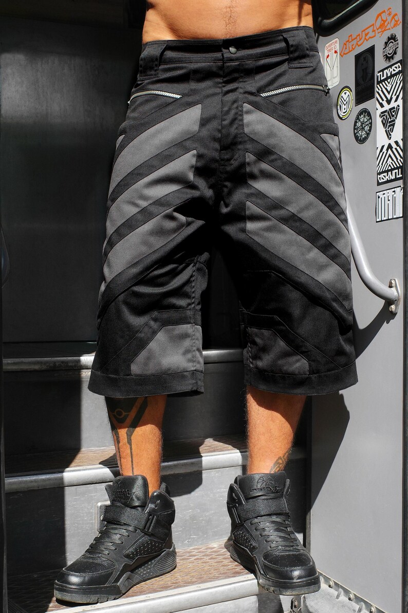 May include: Black shorts featuring dark gray diagonal panels, two horizontal zippered pockets below the waistband, and reinforced stitching. The shorts have a structured design with additional gray paneling near the hem and a button closure.