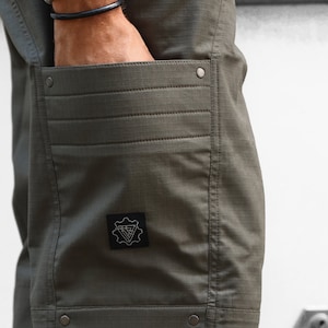 May include: Olive green cargo shorts with a patch on the left thigh featuring a geometric design and the text "V-Gear".