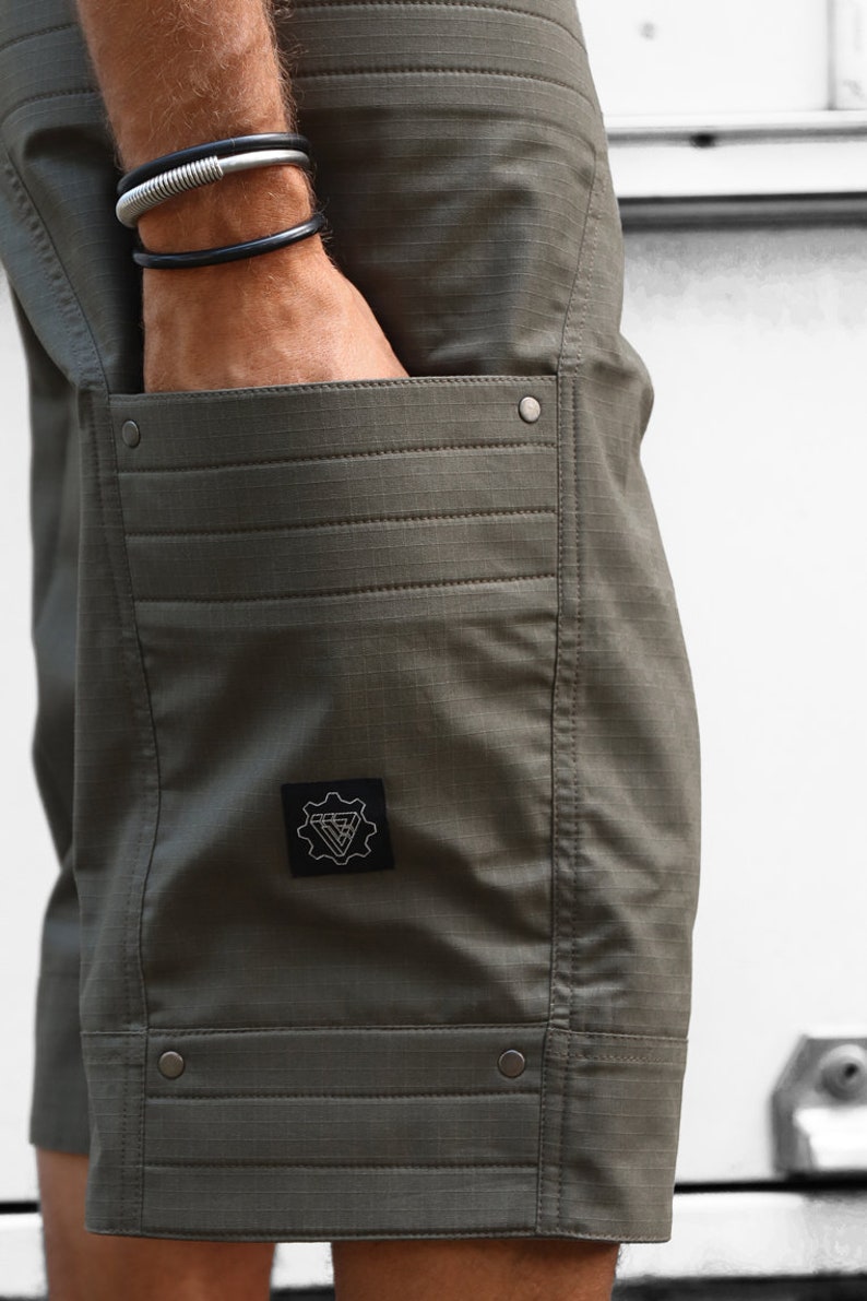 May include: Olive green cargo shorts with a patch on the left thigh featuring a geometric design and the text "V-Gear".