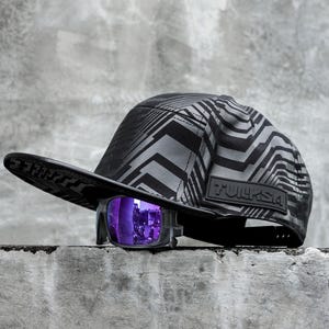 Puede incluir: Una gorra de béisbol negra y gris con estampado, visera negra y la palabra "TURPSA" impresa en el lateral. La gorra está sobre una superficie de hormigón gris con unas gafas de sol moradas delante.