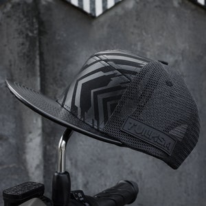 Puede incluir: Una gorra de béisbol negra y gris con estampado y parte trasera de malla. La gorra tiene una visera de cuero negro y un parche negro con la palabra "FULKSA" en ella.