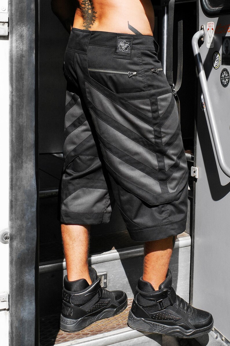 May include: Black cargo shorts featuring a dark gray chevron pattern on the legs, a horizontal zippered pocket on the back, and a small square patch on the waistband. The shorts are paired with black high-top sneakers with textured side panels.