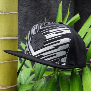 Puede incluir: Una gorra de béisbol negra y plateada con un patrón y un parche hexagonal negro con un logotipo estilizado. La gorra tiene una visera curvada y una corona plana.