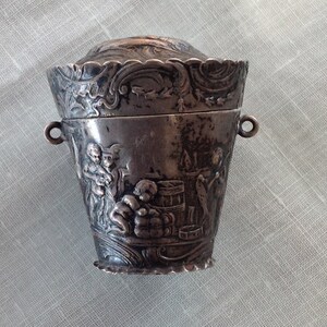 May include: A small, antique silver container with a lid. The container features detailed relief carvings of figures and scenes. It has two small handles and a decorative, scalloped rim. The lid is also decorated with relief details.