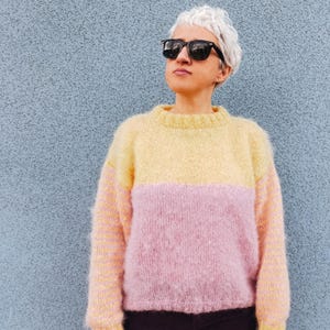 May include: A two-tone jumper in soft yellow and pink. The jumper has a crew neck and long sleeves. The person in the image is wearing black sunglasses and dark purple corduroy trousers. The background is a textured blue wall.