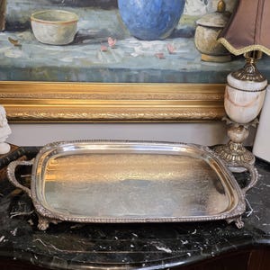 Puede incluir: Una bandeja de servicio rectangular de tono plateado con motivos florales en relieve en la superficie central, un borde decorativo con cuentas, dos asas curvas en los laterales y cuatro patas pequeñas que elevan la base de la bandeja.