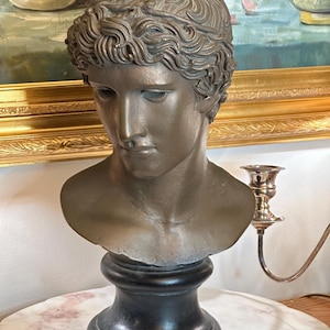 Könnte beinhalten: Bronzefarbene Büste eines jungen Menschen mit lockigem Haar und ruhigem Ausdruck. Die Skulptur ist auf einem schwarzen Sockel im Sockelstil montiert. Die Büste ist ein klassisches Kunstwerk.