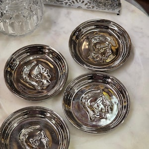 May include: Four silver-colored, round coasters. Each coaster features a raised profile of a Roman figure in a helmet. The coasters are arranged on a white and gray marble surface. Decorative home decor.