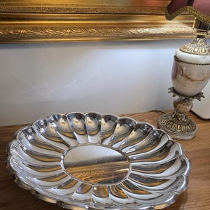 May include: A silver-colored, oval serving dish with a scalloped edge. The dish reflects the surrounding objects, including a lamp and a painting in a gold frame. The dish is on a wooden surface.
