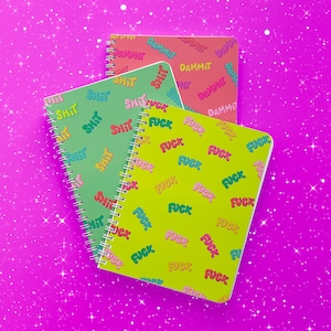 May include: Three spiral bound notebooks with colorful covers. The top notebook has a pink background with the word "DAMMIT" repeated in a rainbow pattern. The middle notebook has a green background with the word "SHIT" repeated in a rainbow pattern. The bottom notebook has a yellow background with the word "FUCK" repeated in a rainbow pattern.