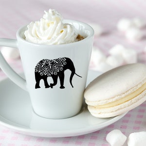 May include: A white ceramic coffee cup with a black silhouette of an elephant with a mandala design on its back. The cup is filled with coffee and topped with whipped cream. A white macaron is on a white saucer next to the cup.