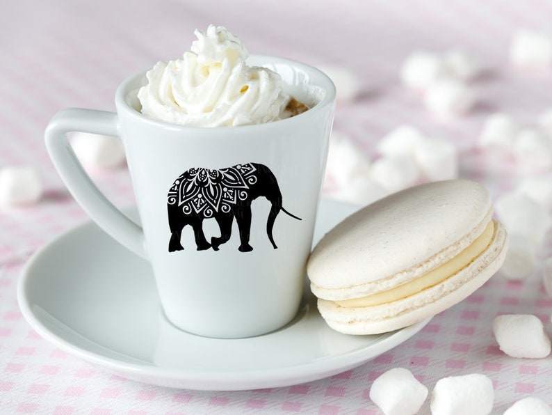 May include: A white ceramic coffee cup with a black silhouette of an elephant with a mandala design on its back. The cup is filled with coffee and topped with whipped cream. A white macaron is on a white saucer next to the cup.