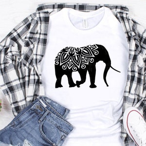 May include: A white t-shirt with a black silhouette of an elephant with a mandala design on its head. The elephant is facing right and has a long trunk. The t-shirt is laying on a plaid shirt and a pair of blue jeans.