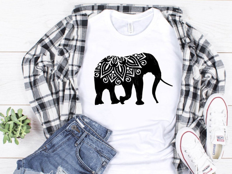 May include: A white t-shirt with a black silhouette of an elephant with a mandala design on its head. The elephant is facing right and has a long trunk. The t-shirt is laying on a plaid shirt and a pair of blue jeans.