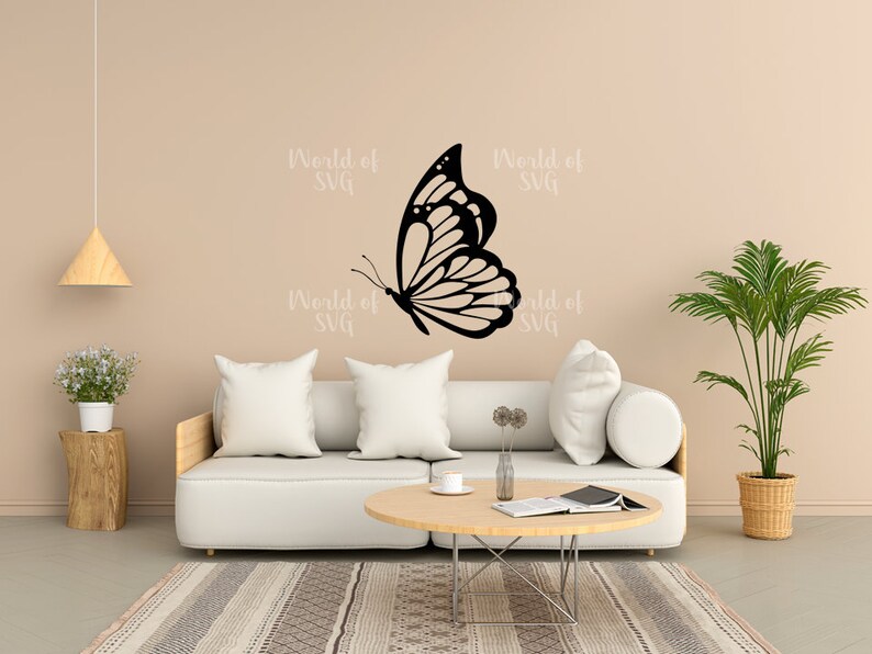 May include: A white couch with two pillows and a coffee table in front of it. A black butterfly silhouette is on the wall behind the couch. The wall is a light beige color. There is a potted plant in the corner of the room.
