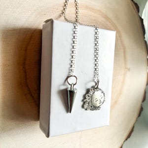 May include: Two silver dowsing pendulums, one shaped like a cone and the other with a sun face design, hang from silver chains.